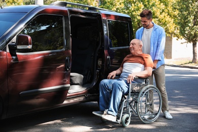Photo of Young man helping patient in wheelchair to get into van outdoors