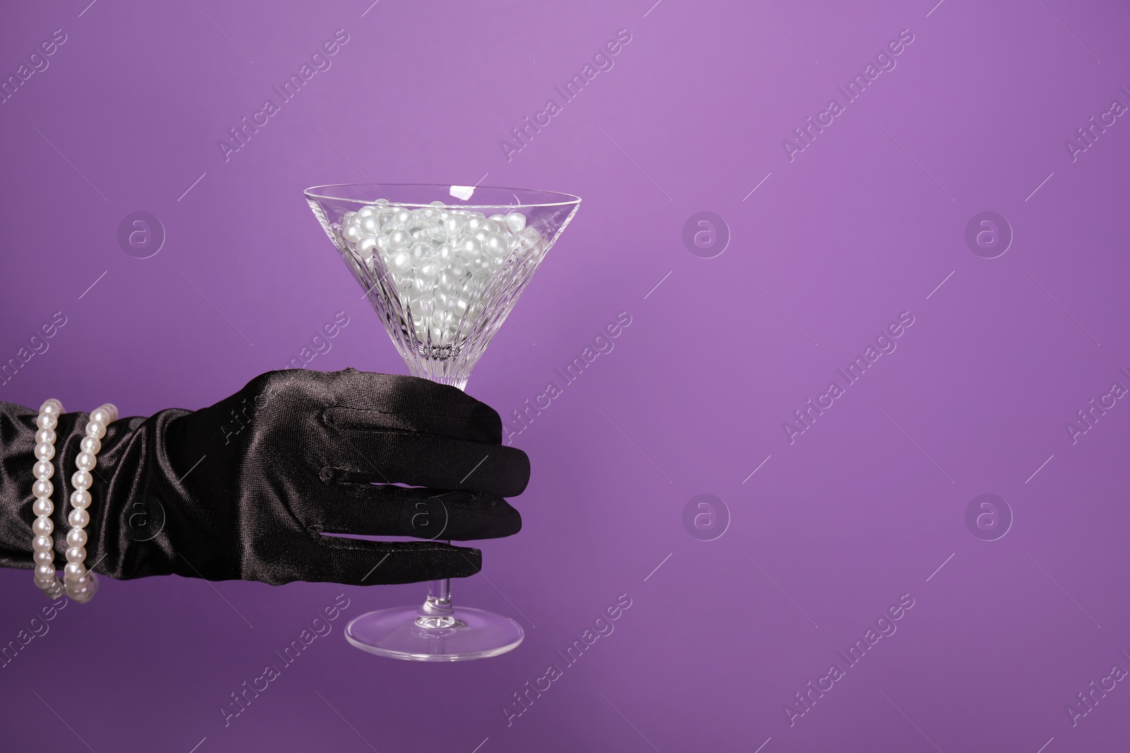 Photo of Woman in elegant gloves holding beautiful martini glass with pearls on purple background, closeup. Space for text