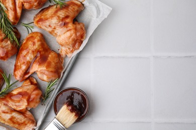 Photo of Raw chicken wings, rosemary, marinade and basting brush on light tiled table, flat lay. Space for text