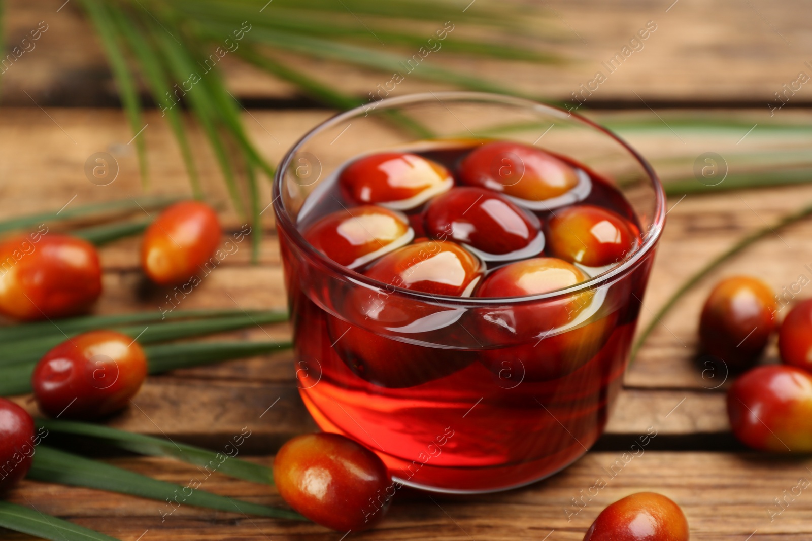 Photo of Palm oil in glass with fruits and tropical leaves on wooden table, closeup