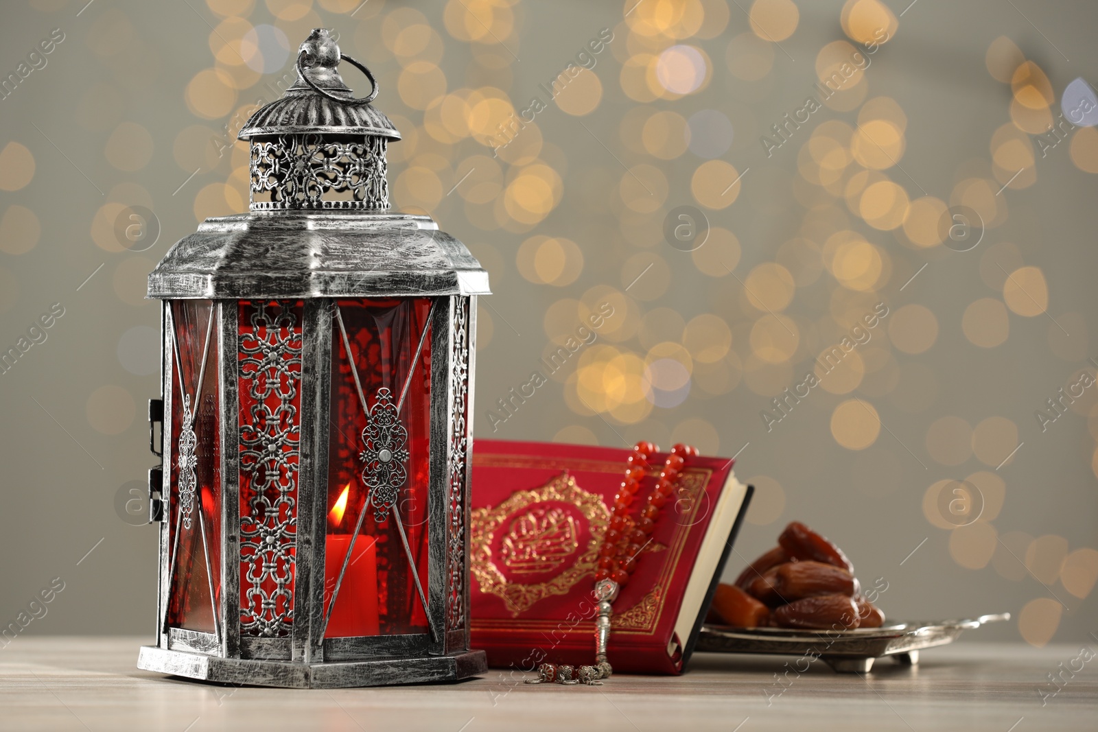 Photo of Arabic lantern, Quran, misbaha and dates on table against blurred lights