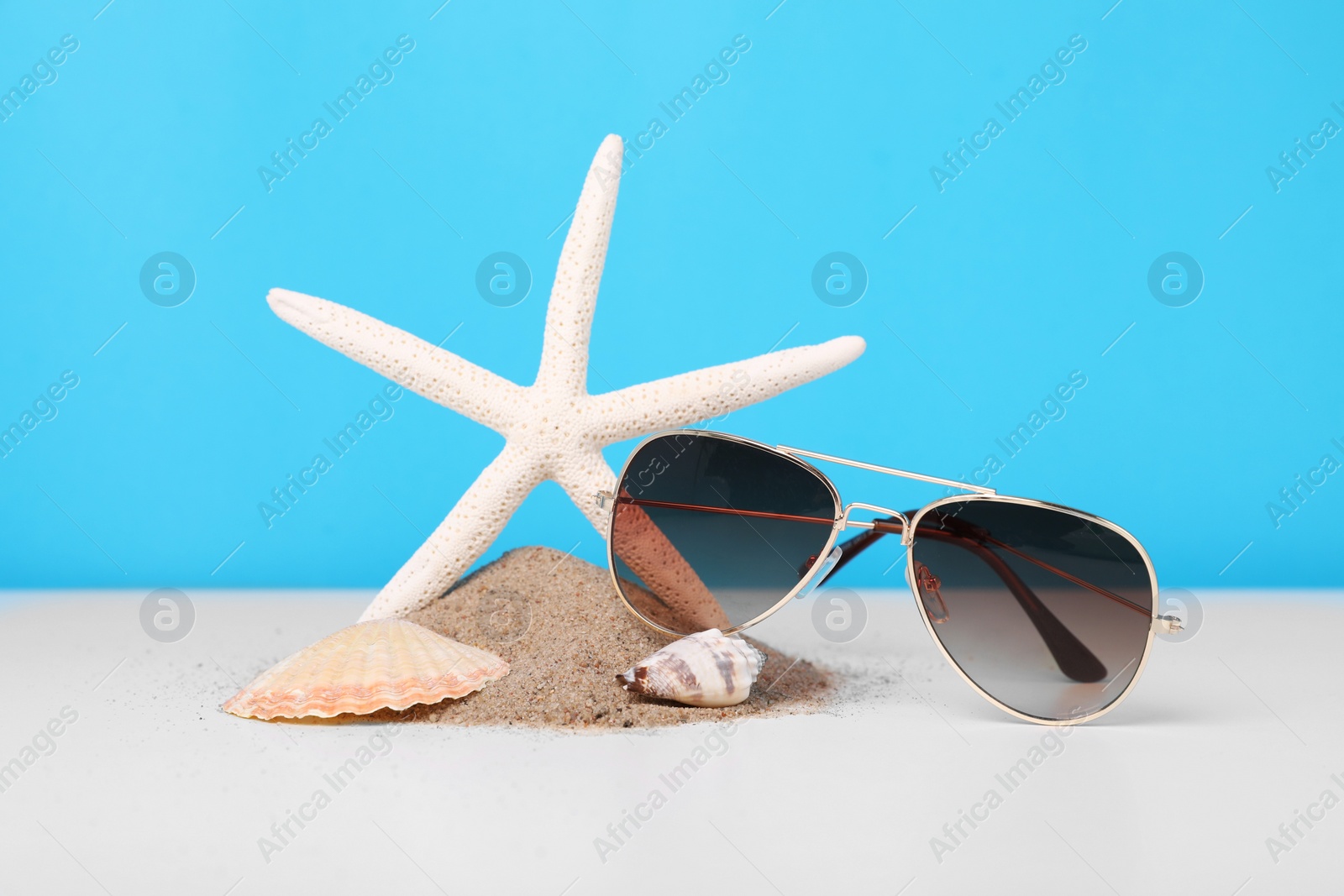 Photo of Stylish sunglasses, seashells, starfish and sand on white table against light blue background