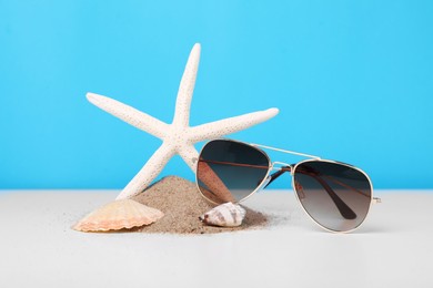 Stylish sunglasses, seashells, starfish and sand on white table against light blue background