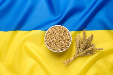 Photo of Bowl of wheat grains and ears on national Ukrainian flag, flat lay