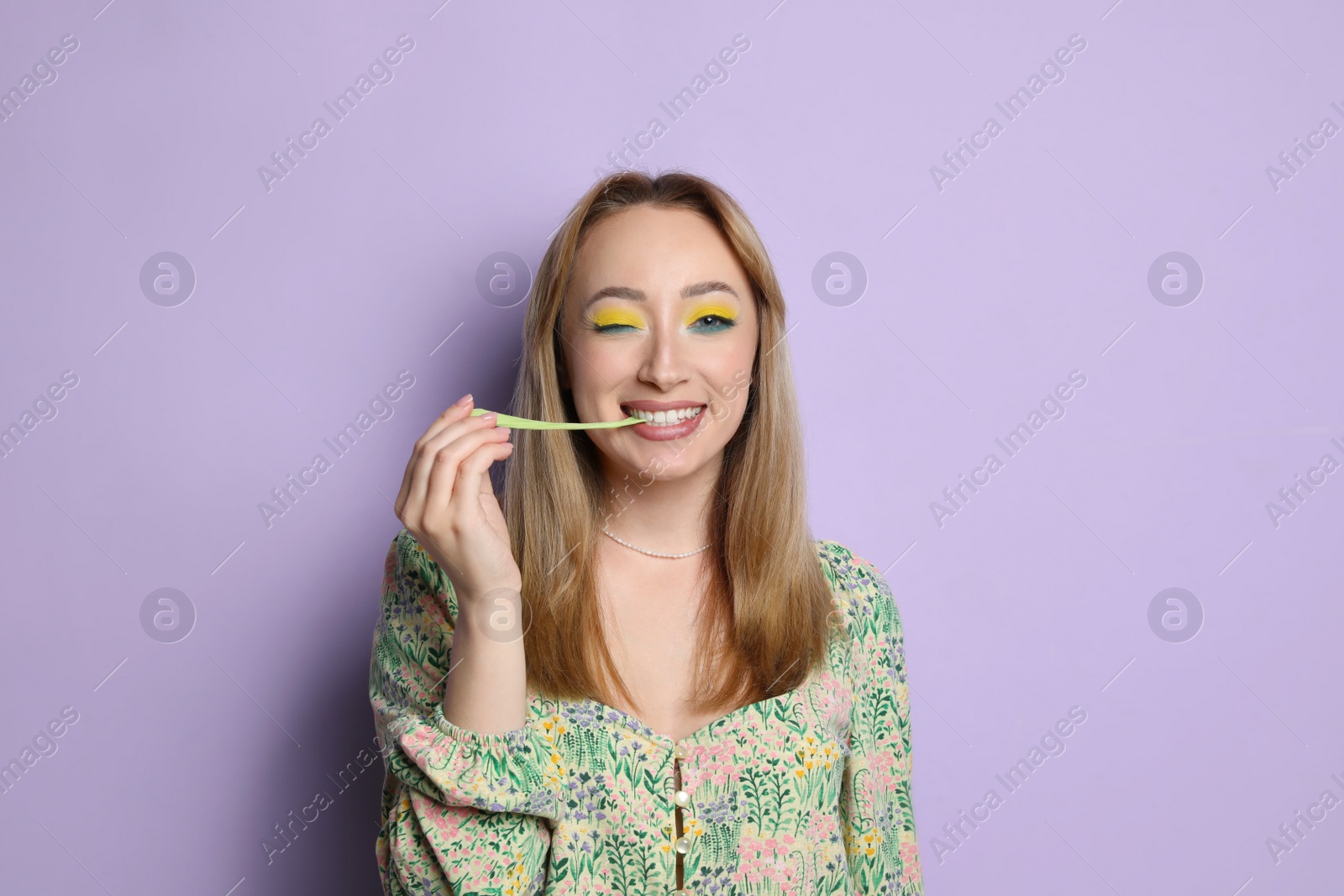 Photo of Fashionable young woman with bright makeup chewing bubblegum on lilac background