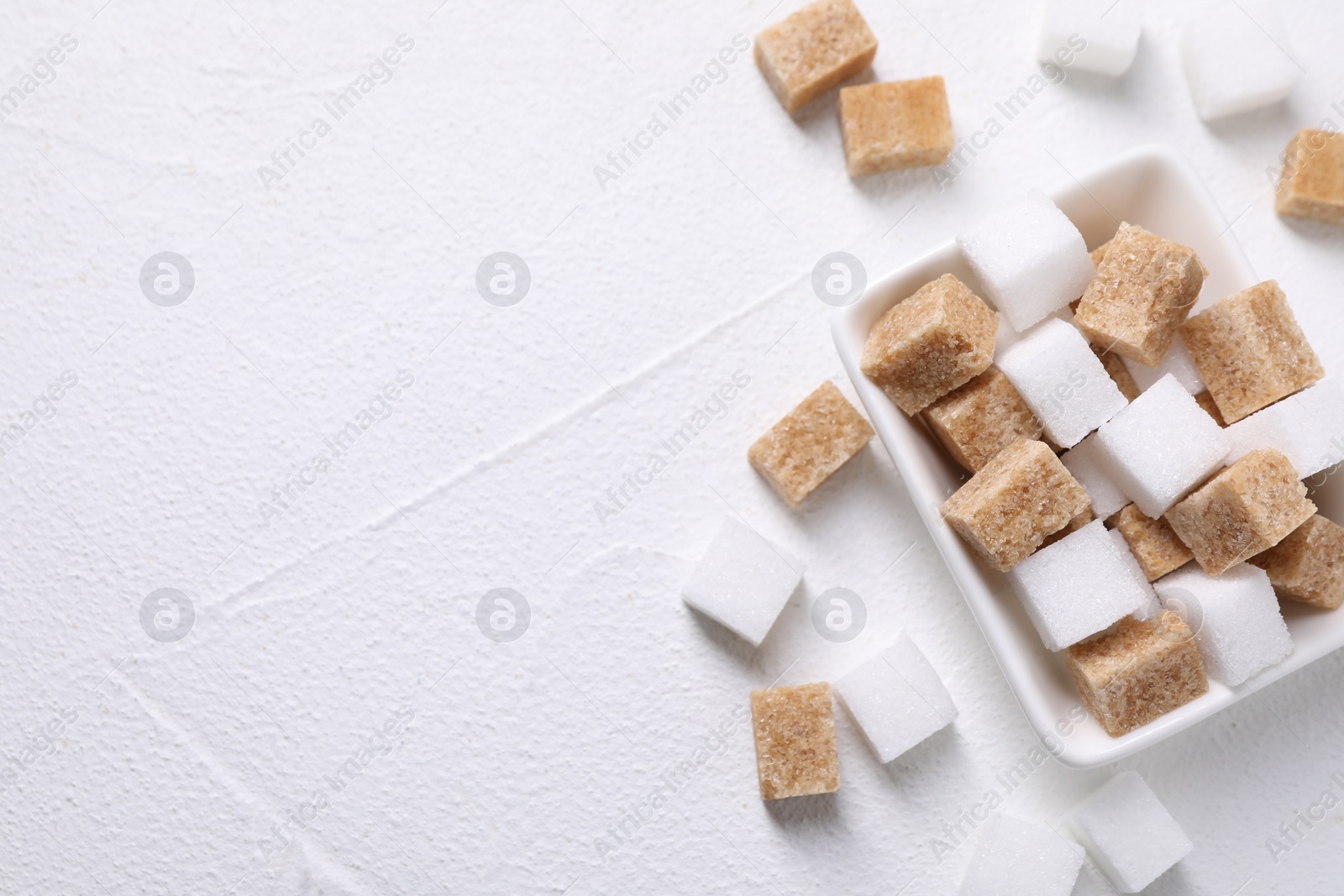 Photo of Different sugar cubes in bowl on white table, top view. Space for text