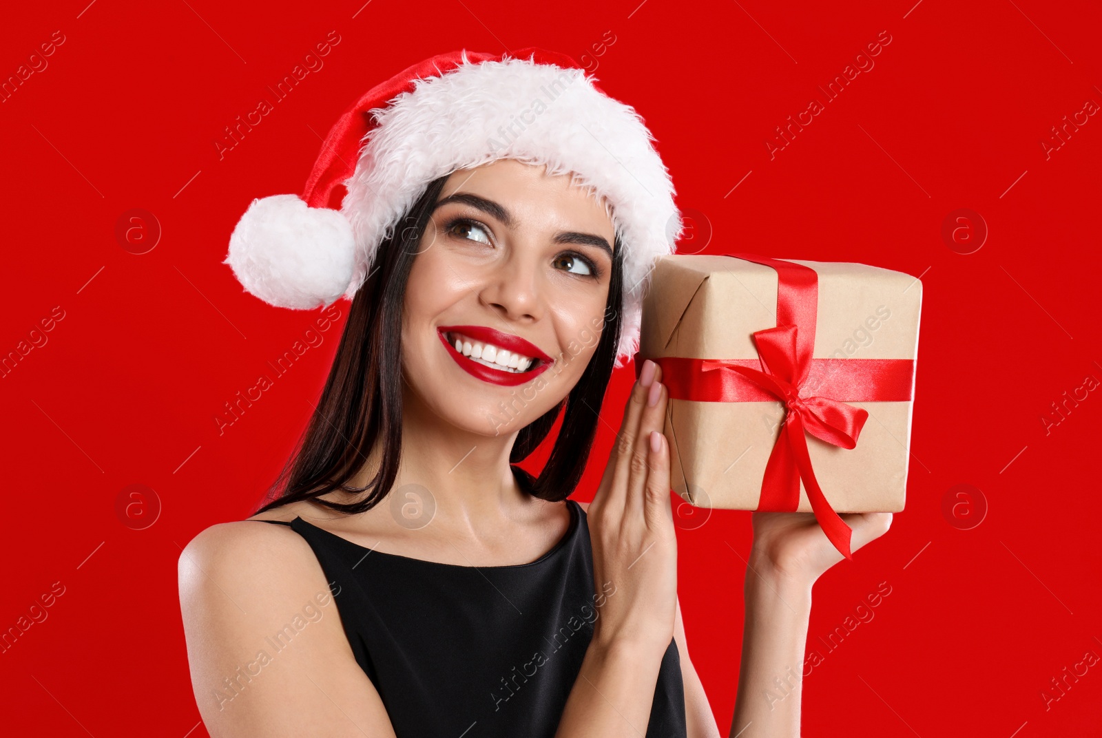 Photo of Woman in black dress and Santa hat holding Christmas gift on red background