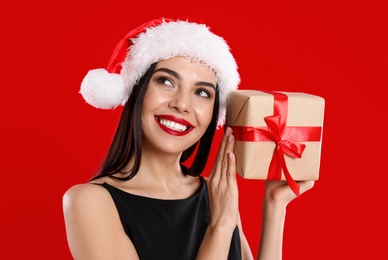 Woman in black dress and Santa hat holding Christmas gift on red background