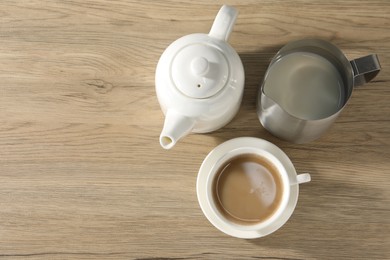 Photo of Aromatic tea with milk in cup, teapot and pitcher on wooden table, flat lay. Space for text