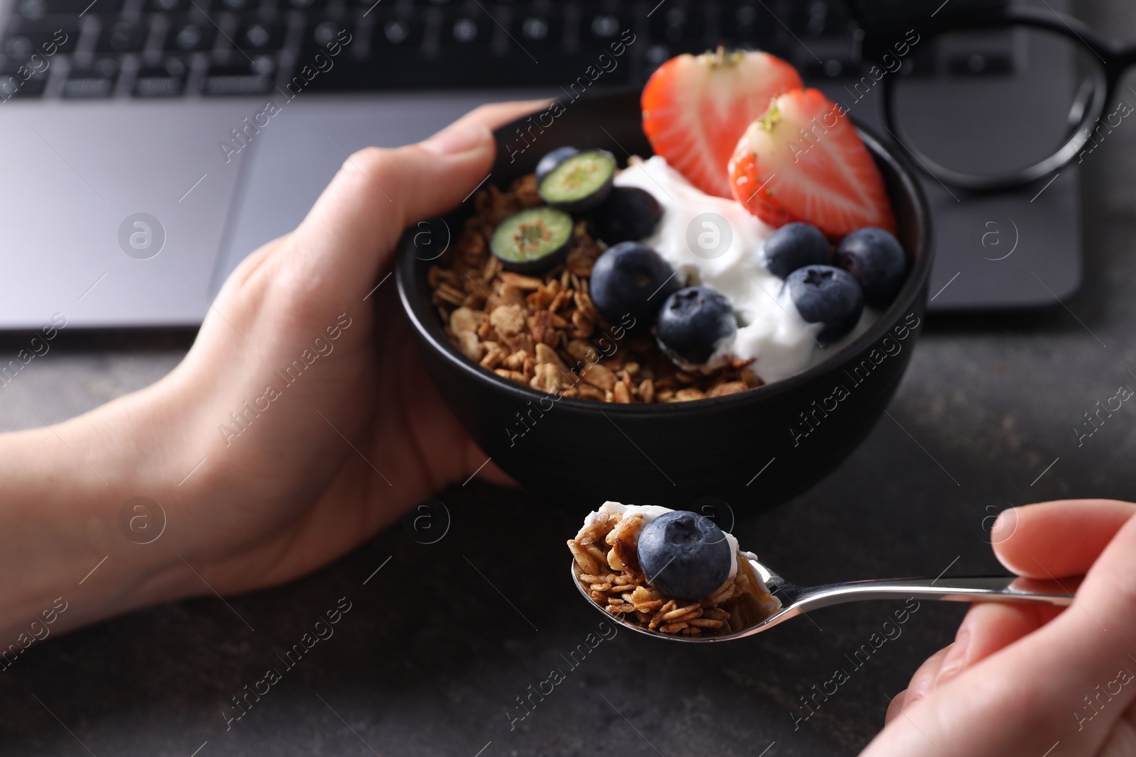 Photo of Woman eating tasty granola with yogurt and berries at workplace, closeup