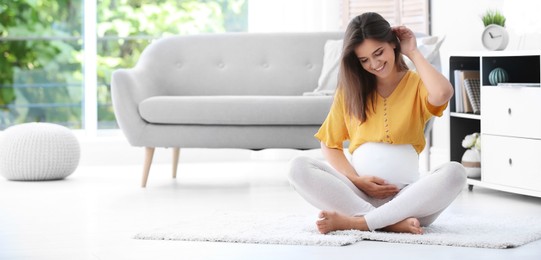 Happy pregnant woman sitting on floor at home, space for text. Banner design