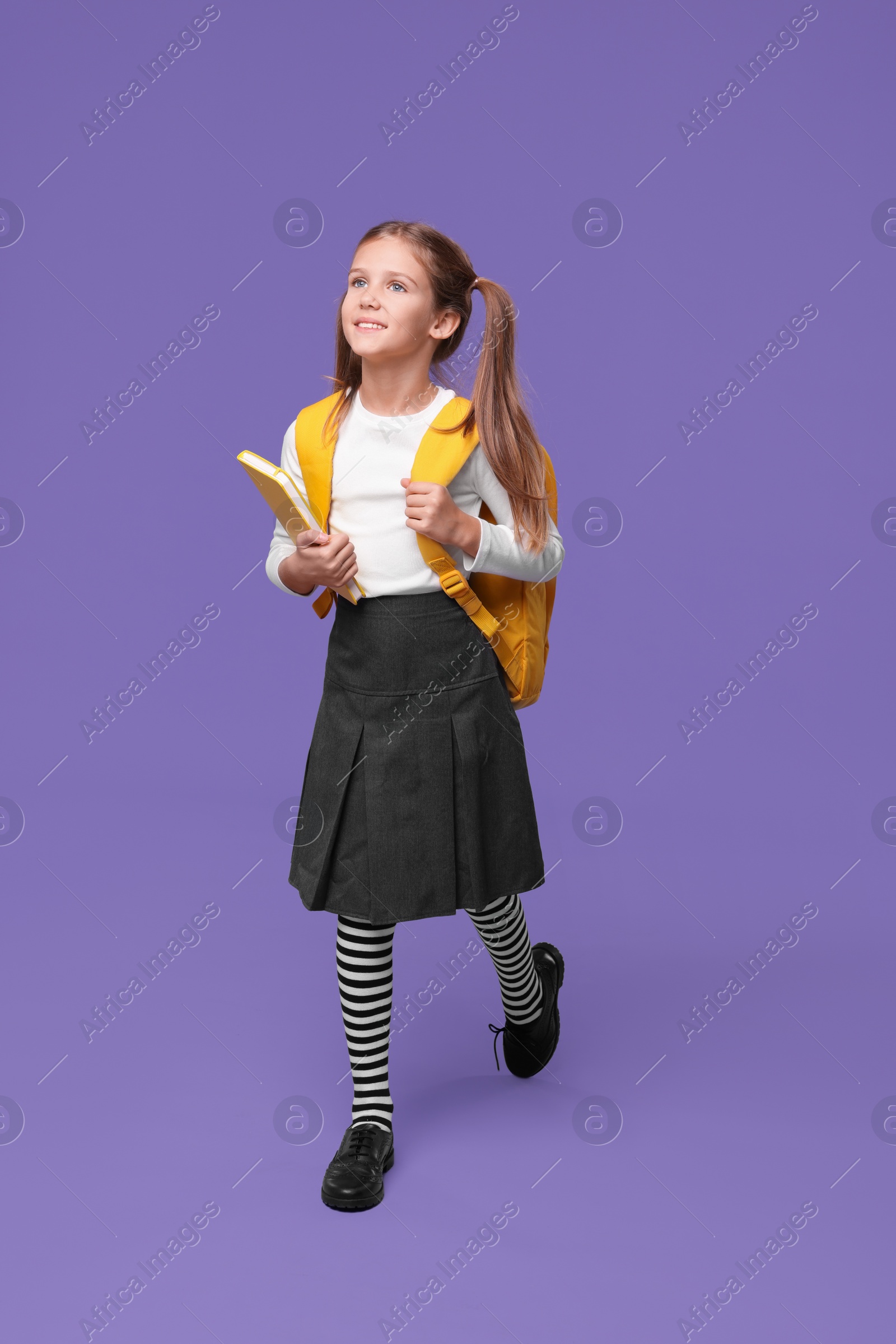 Photo of Smiling schoolgirl with backpack and book on violet background