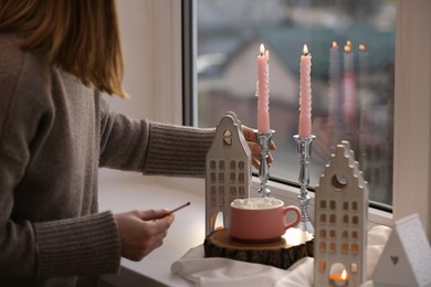 Photo of Woman taking candle from windowsill with house shaped lanterns indoors, closeup