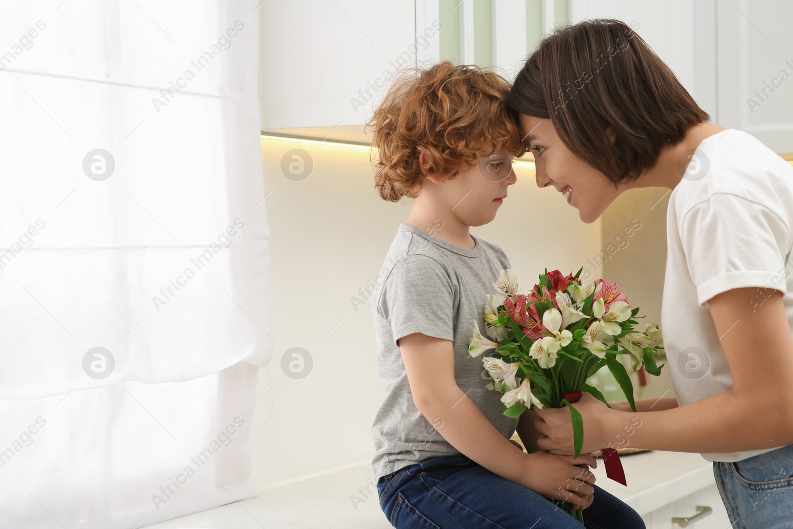 Photo of Happy woman with her cute son and bouquet of beautiful flowers in kitchen, space for text. Mother's day celebration