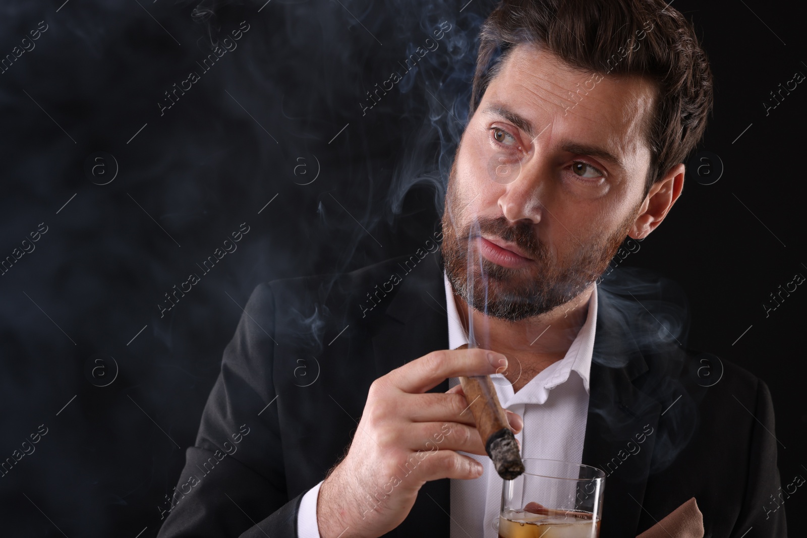 Photo of Handsome man in elegant suit with glass of whiskey smoking cigar on black background. Space for text