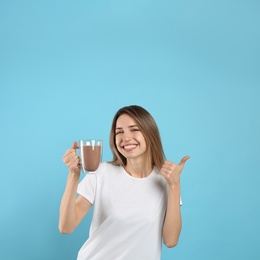 Young woman with glass cup of chocolate milk showing thumb up on light blue background