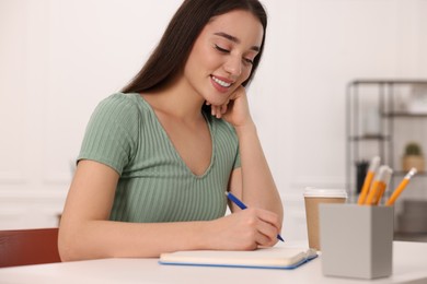 Photo of Young woman writing in notebook at white table indoors