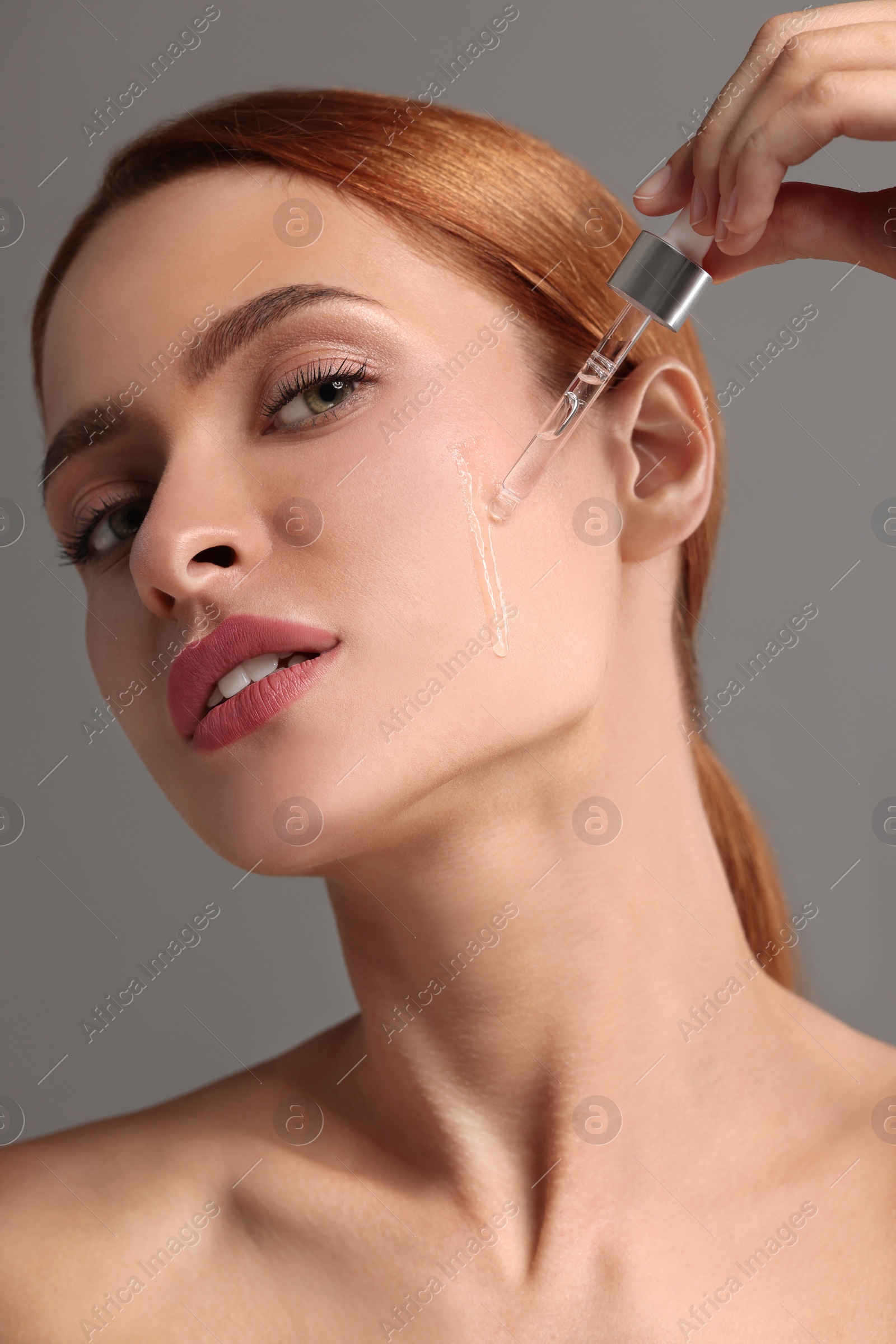 Photo of Beautiful young woman applying cosmetic serum onto her face on grey background, closeup