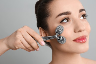Woman using metal face roller on grey background, closeup