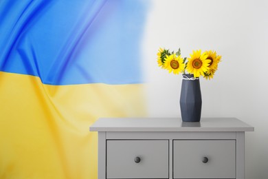 Bouquet of beautiful sunflowers in vase on commode near light wall with Ukrainian national flag