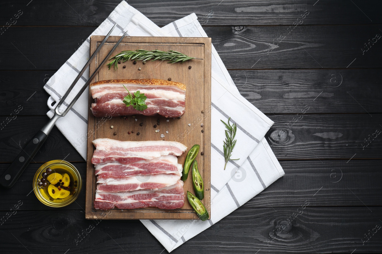 Photo of Pieces of raw pork belly, chili pepper, peppercorns, rosemary and parsley on black wooden table, flat lay