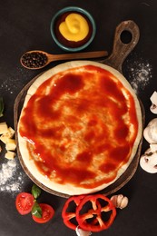 Photo of Pizza dough smeared with tomato sauce and products on dark table, flat lay