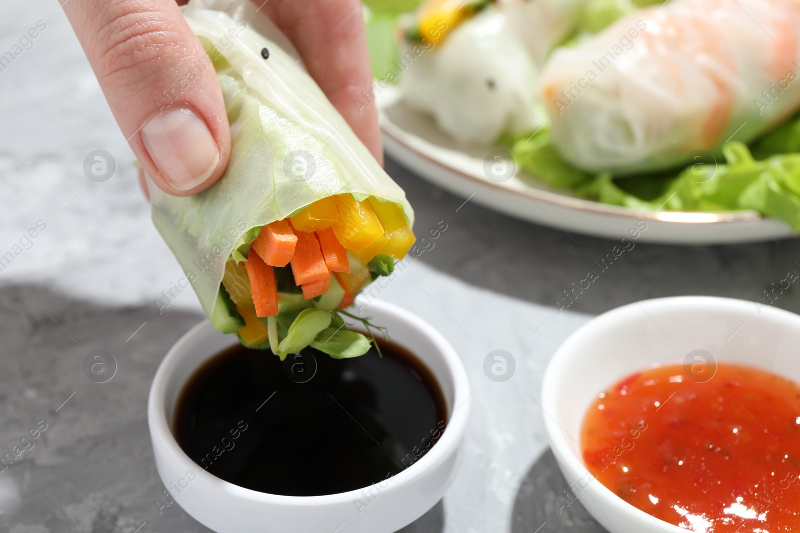 Photo of Woman dipping delicious spring roll into soy sauce at grey table, closeup