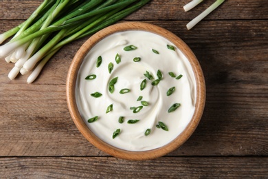 Photo of Bowl of fresh sour cream with green onion on wooden table, flat lay