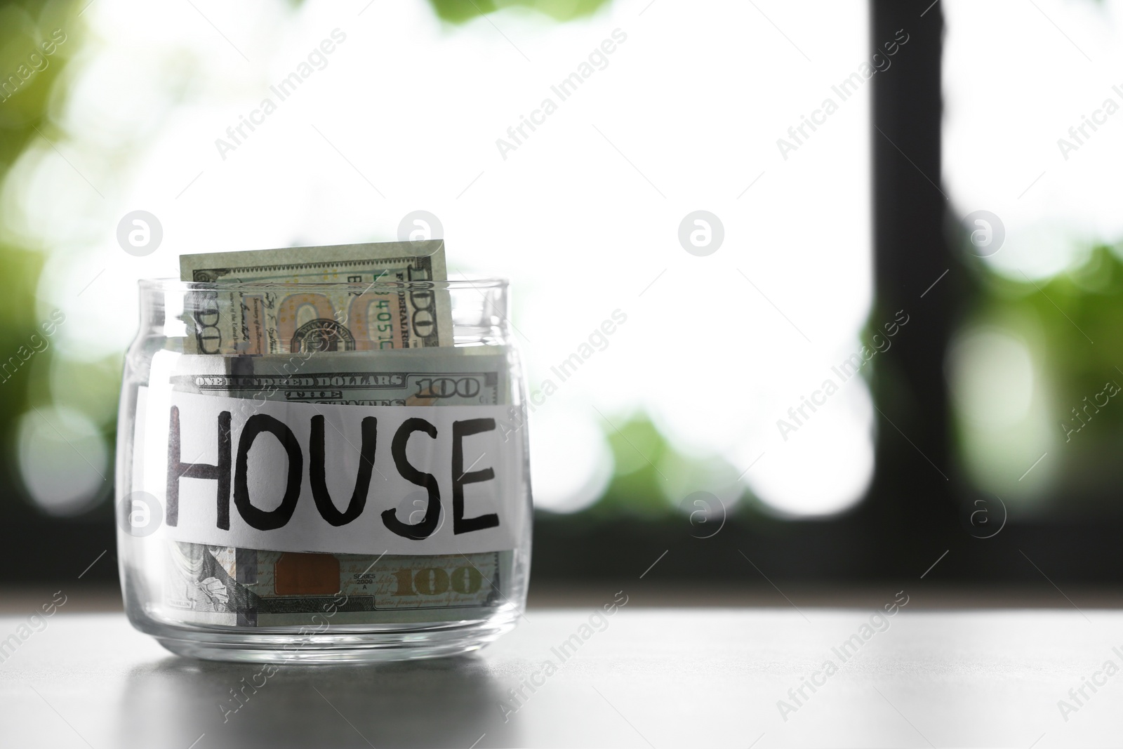Photo of Glass jar with money and word HOUSE on table against blurred background, space for text