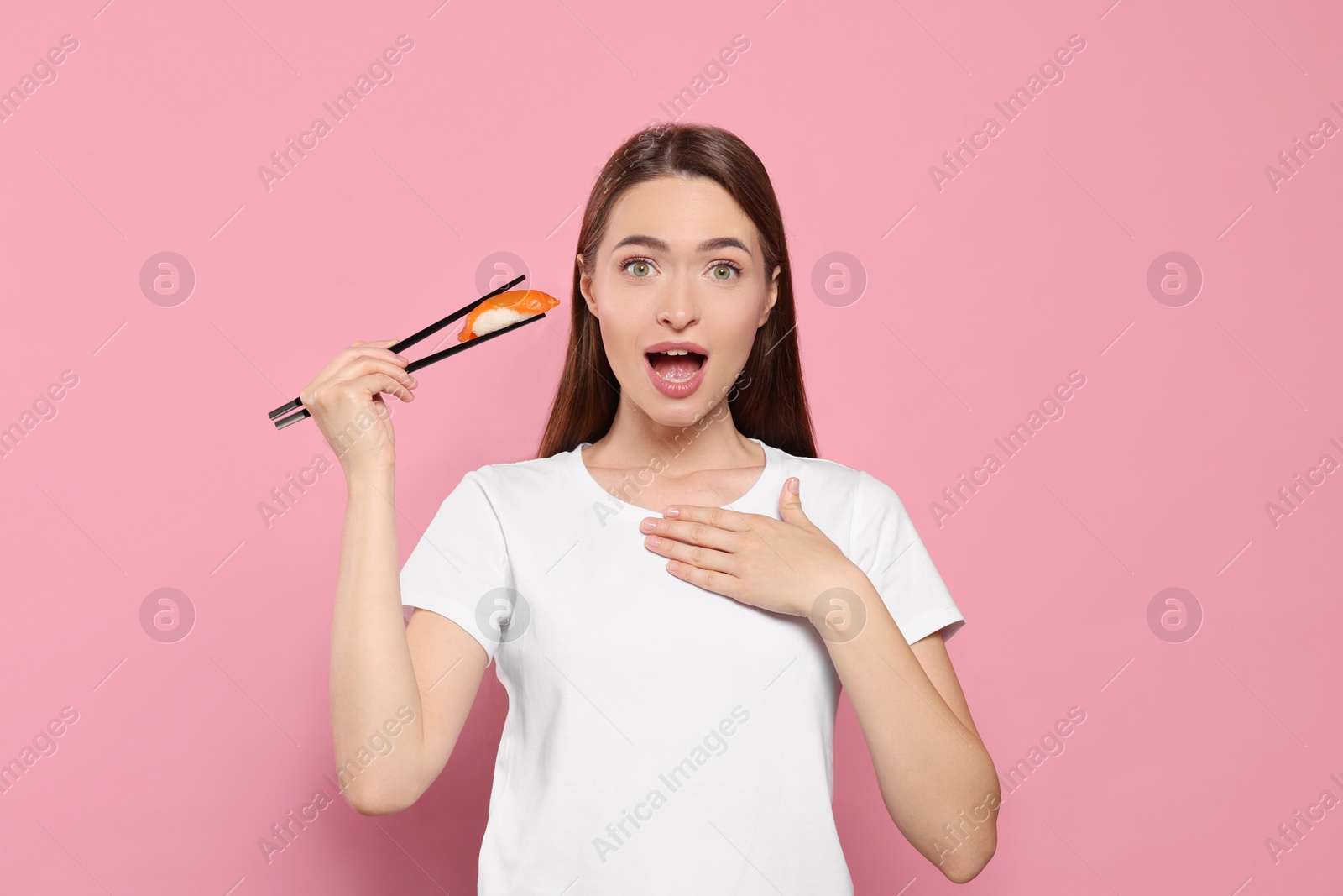 Photo of Emotional young woman holding sushi with chopsticks on pink background