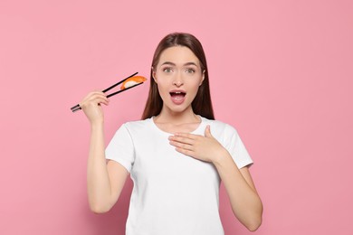 Emotional young woman holding sushi with chopsticks on pink background