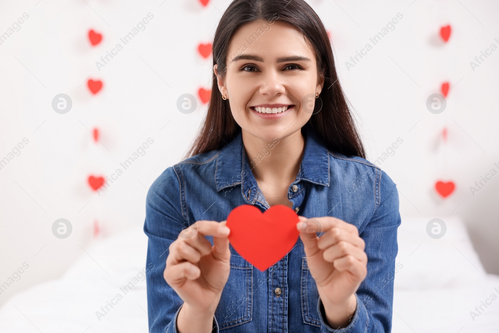 Photo of Beautiful young woman with decorative red heart indoors, view from camera. Valentine's day celebration in long distance relationship