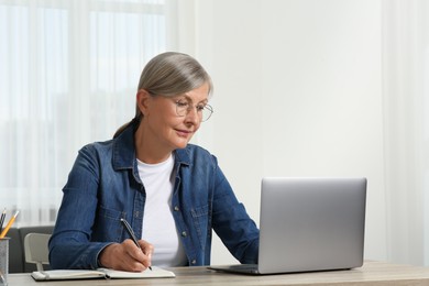Photo of Beautiful senior woman writing notes in notebook while using laptop at wooden table indoors