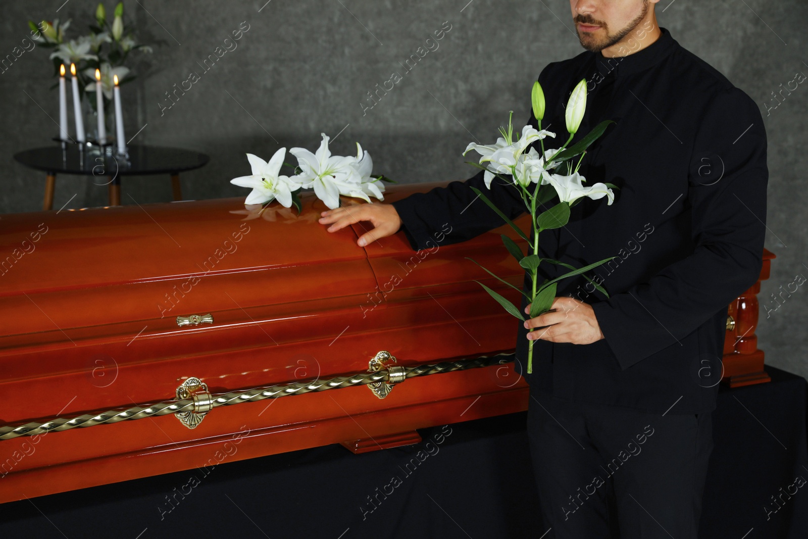 Photo of Young man with white lilies near casket in funeral home, closeup