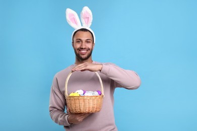 Happy African American man in bunny ears headband holding wicker basket with Easter eggs on light blue background
