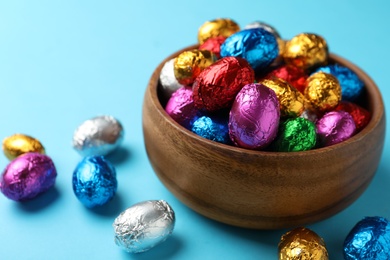 Photo of Wooden bowl with chocolate eggs wrapped in colorful foil on light blue background