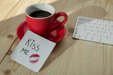 Sticky note with phrase Kiss Me, lipstick mark, cup of coffee and keyboard on wooden table