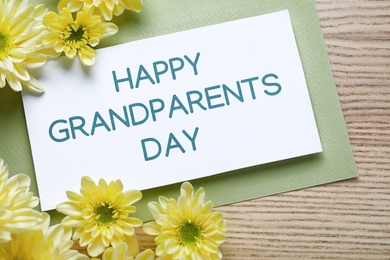 Photo of Beautiful yellow flowers and card with phrase Happy Grandparents Day on wooden background, flat lay