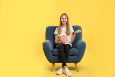 Teenage girl with popcorn sitting in armchair during cinema show on color background