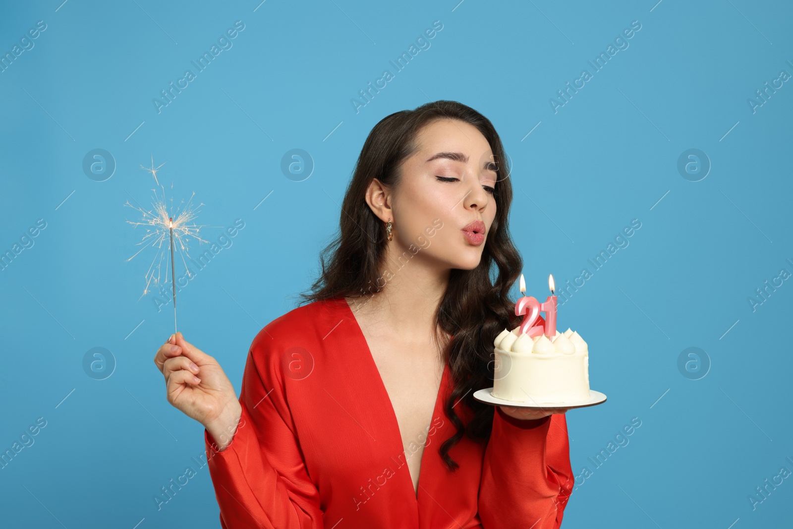 Photo of Coming of age party - 21st birthday. Woman holding sparkler and blowing number shaped candles on cake against light blue background