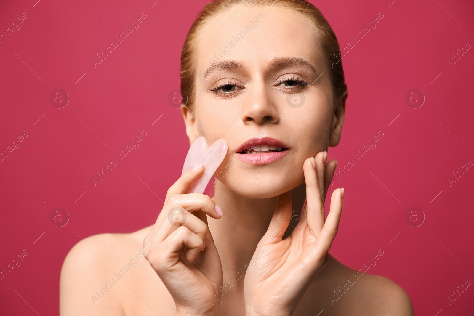 Photo of Beautiful young woman doing facial massage with gua sha tool on pink background, closeup