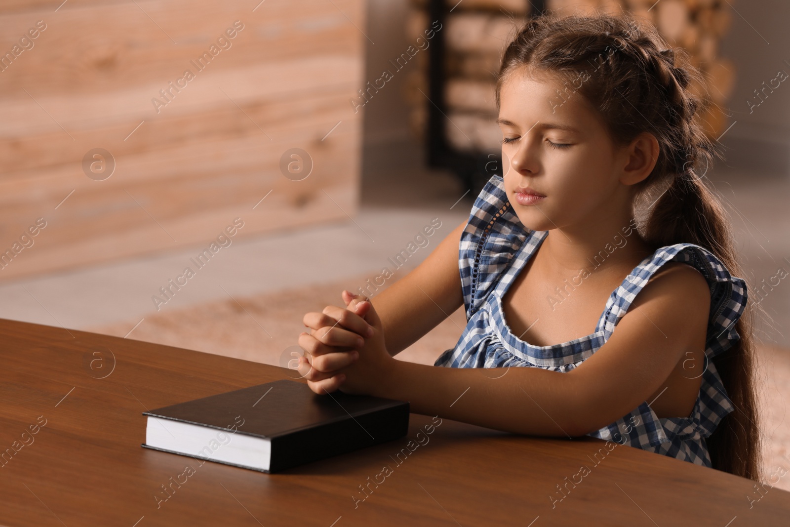 Photo of Cute little girl praying over Bible at table in room