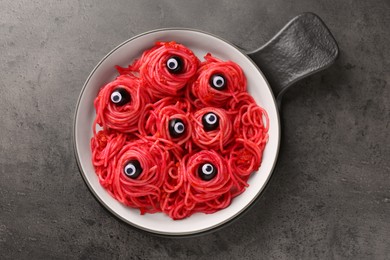 Photo of Red pasta with decorative eyes and olives in bowl on grey textured table, top view. Halloween food