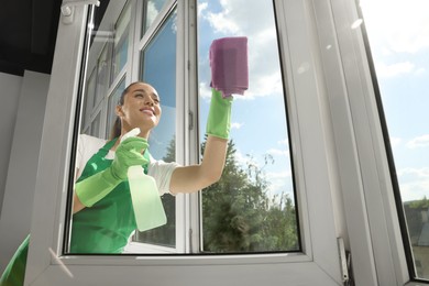 Photo of Happy young woman cleaning window glass with rag and spray indoors, low angle view