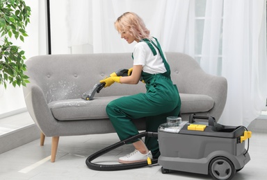 Female janitor removing dirt from sofa with upholstery cleaner in room