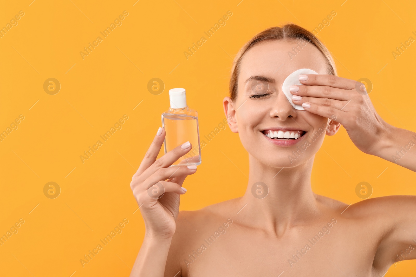 Photo of Smiling woman removing makeup with cotton pad and holding bottle on yellow background. Space for text