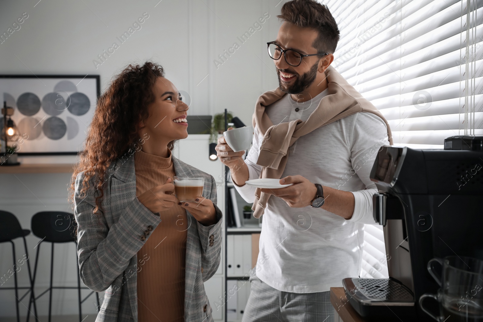 Photo of Colleagues with hot drinks talking near modern coffee machine in office