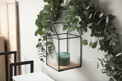 Beautiful garland made of eucalyptus branches and aromatic candle hanging on white wall indoors