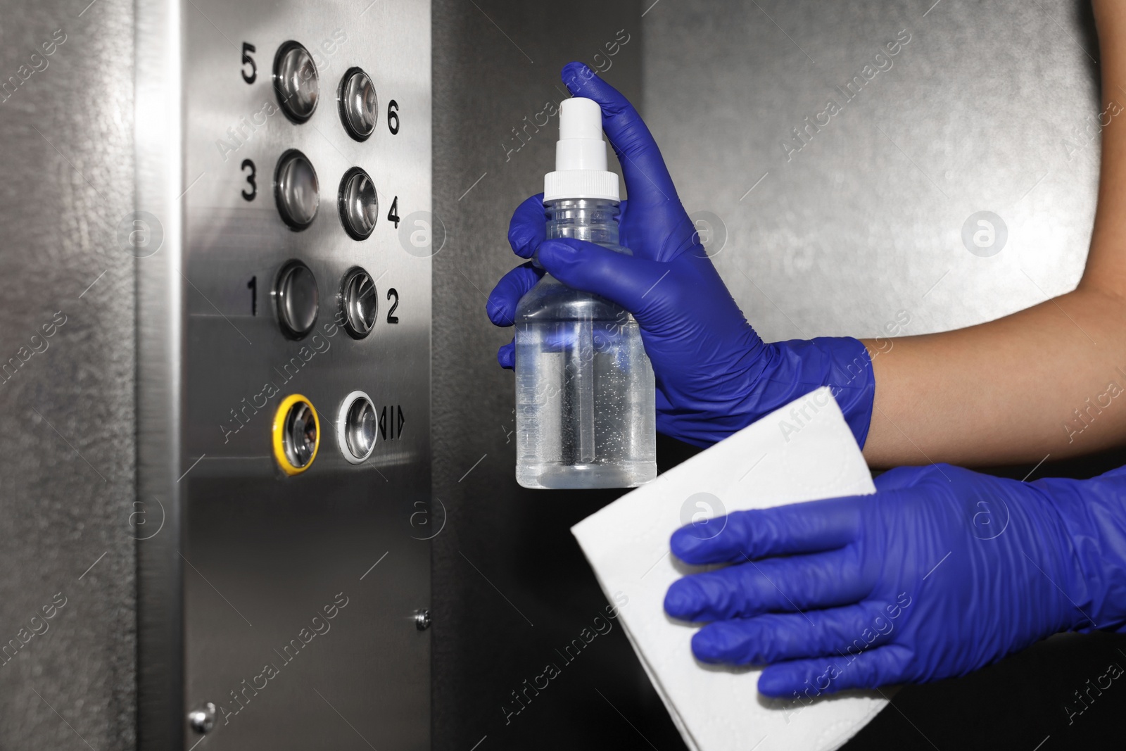 Photo of Woman wiping buttons of elevator`s panel with detergent and paper napkin, closeup