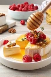 Photo of Pouring honey onto brie cheese served with raspberries and walnuts on white wooden table, closeup